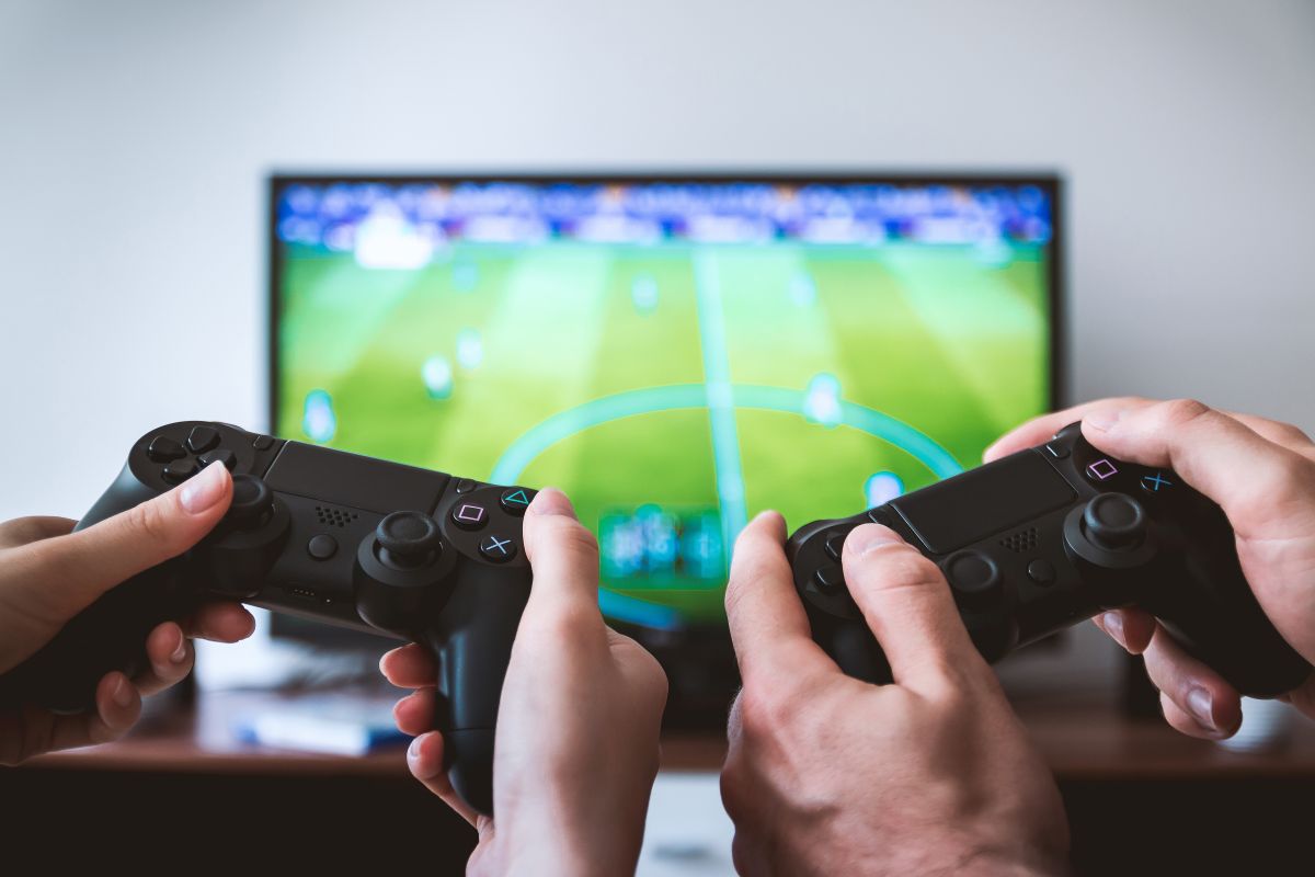 Duas pessoas jogando com controles diante da TV, com uma partida de futebol na tela